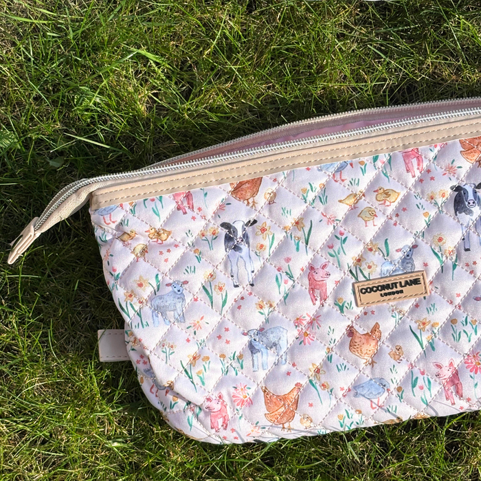 Quilted Coconut Lane expandable makeup bag with farmyard animal print on green grass in sunlight.