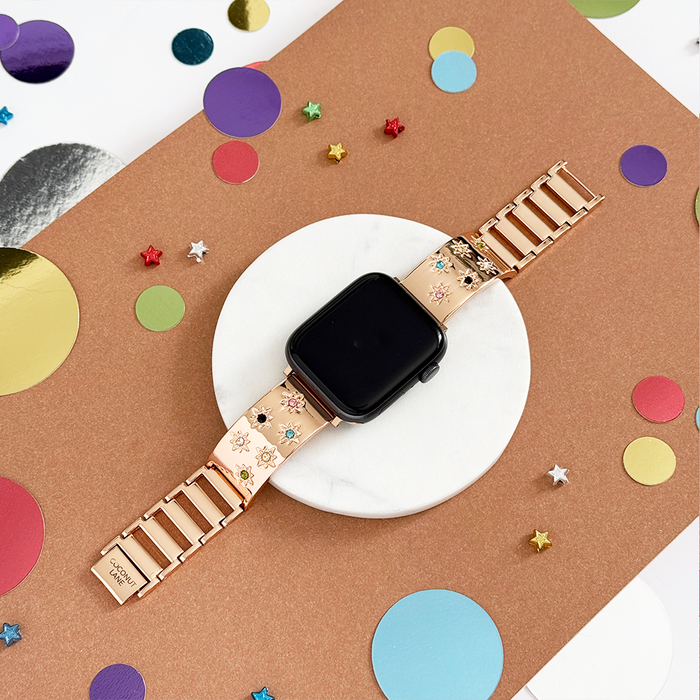Rose gold Apple Watch strap with colored stones on a brown and white background decorated with confetti and stars.
