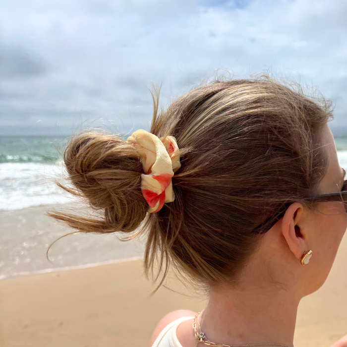 Woman wearing sunglasses and a Coconut Lane floral scrunchie stands on the beach, facing the ocean.