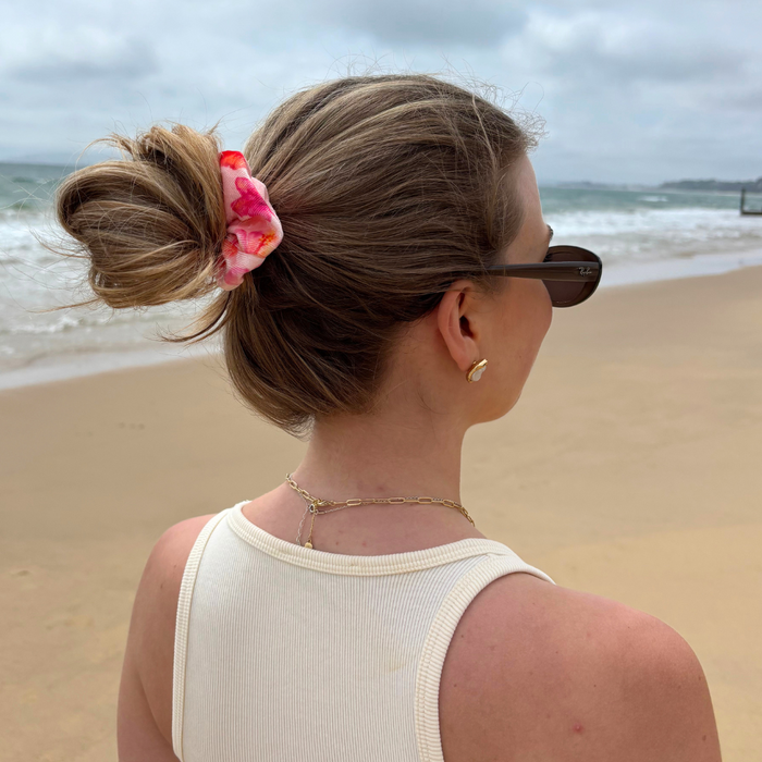 Woman with a messy bun wears a pink hibiscus-patterned scrunchie on a sandy beach, facing the ocean.