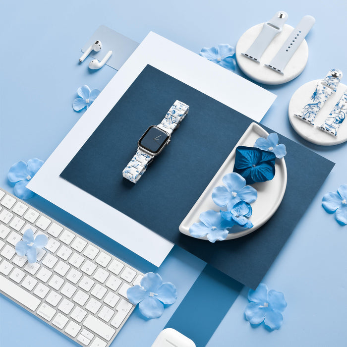 A flat lay photo featuring a Coconut Lane Luxe Blue & White Apple Watch Strap on blue and white paper backgrounds. Surrounding items include a keyboard, AirPods, ceramic holders, and blue flower petals arranged artistically. The color theme is predominantly blue and white.