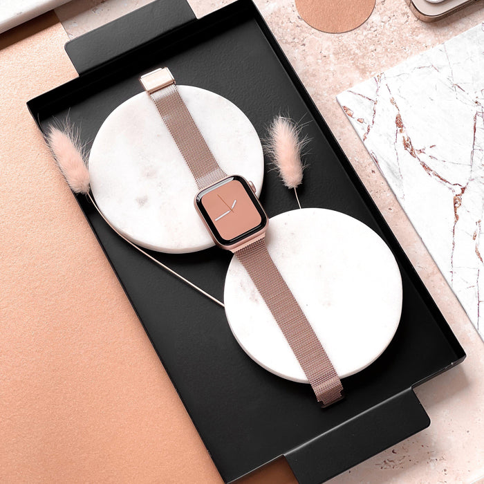 A rose gold smartwatch, featuring the Milanese Apple Watch Strap - Rose Gold by Coconut Lane, is neatly placed on two white circular bases within a black tray. Minimalist decorations, including beige feathers and marble-patterned items, surround the tray on a blush-toned surface.