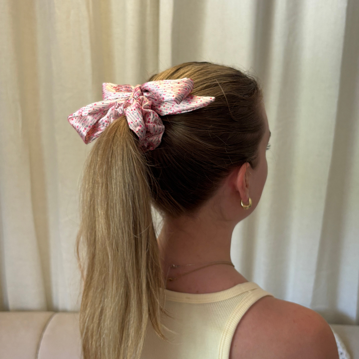 Blonde woman with a ponytail wearing a pink satin bow scrunchie, seen from behind.