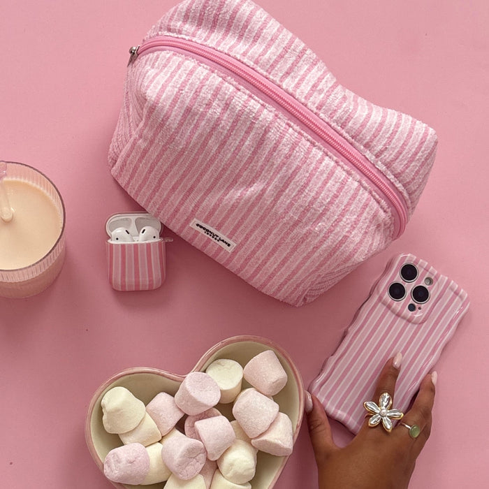 Powder striped towel makeup bag by Coconut Lane with a phone, AirPods case, candle and heart marshmallows on pink surface.