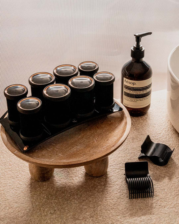 Set of hair rollers on a wooden stand with Aesop bottle and comb on a neutral background
