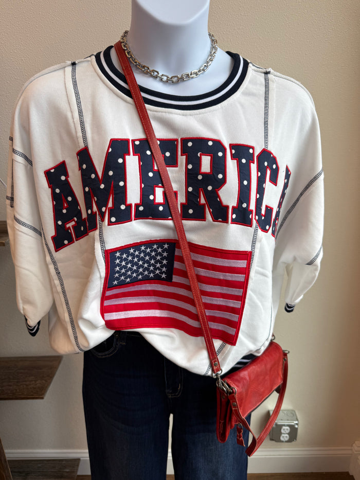 Cream short-sleeve sweatshirt with bold red-and-navy text and small American flag patch