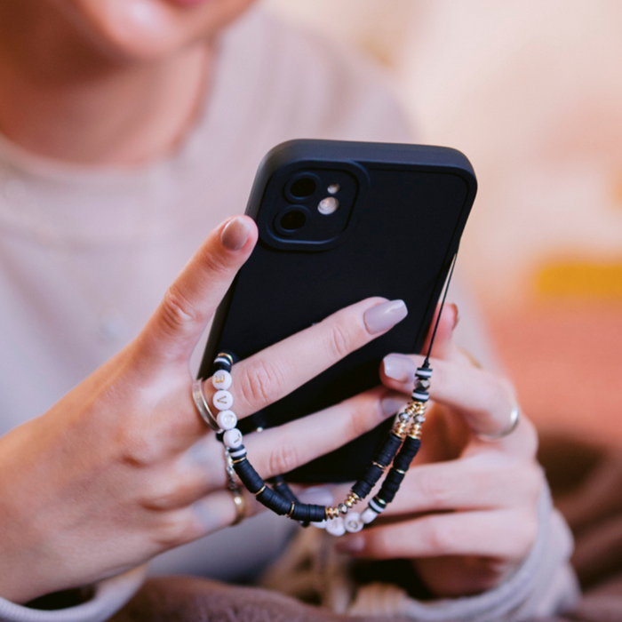 Hand holding smartphone with Coconut Lane beaded phone strap in black and gold; person smiling in background.