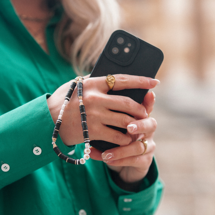Black smartphone with Coconut Lane black and gold beaded phone strap held by woman in green shirt wearing gold rings.