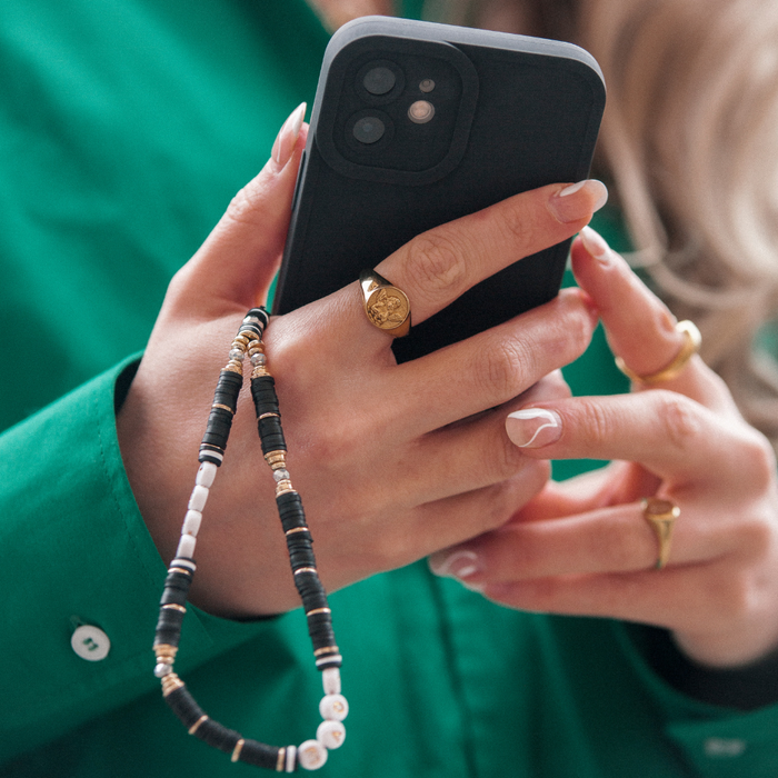 Coconut Lane beaded phone strap in black and gold on a phone held by someone wearing a green shirt and gold rings.