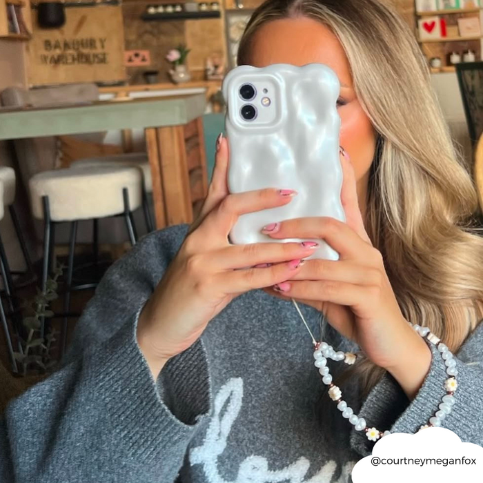 Woman with wavy blonde hair takes a selfie in a cafe, holding a phone with a beaded daisy pearl strap.