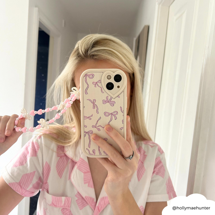 Woman in pink and white pajamas holds phone with Coconut Lane Hearts & Bows beaded strap, taking a mirror selfie.