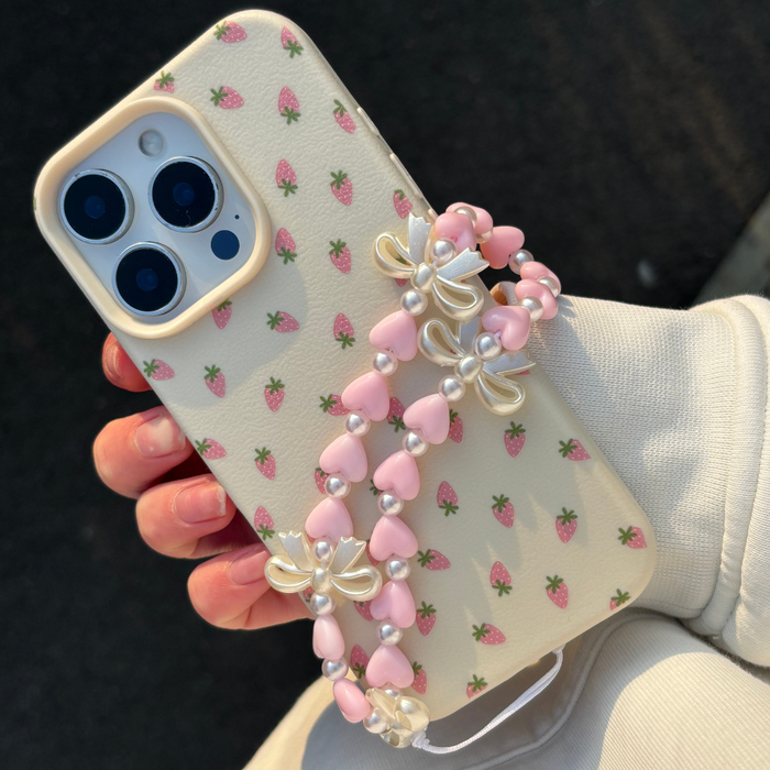 Hand holding a phone with a red strawberry case and Coconut Lane beaded strap featuring hearts and bows.
