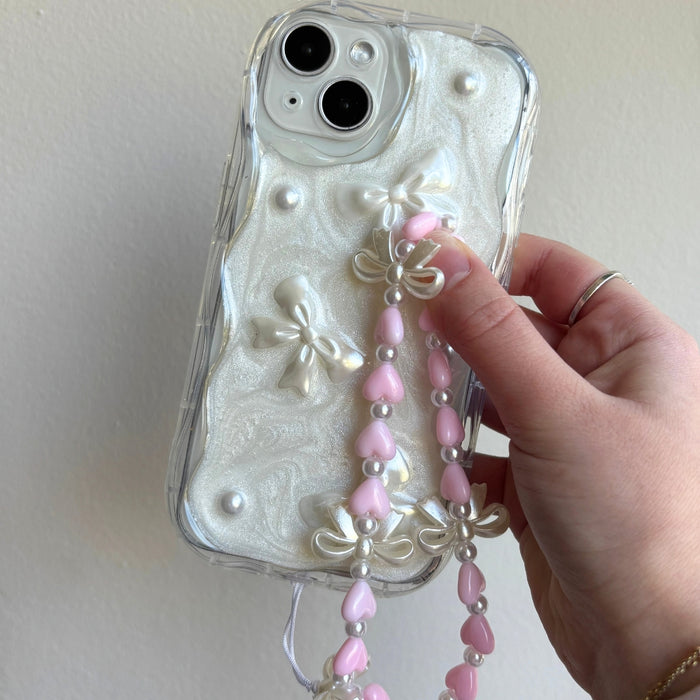 Hand holding a phone with a beaded strap featuring hearts and bows, in a pearly case decorated with bows.
