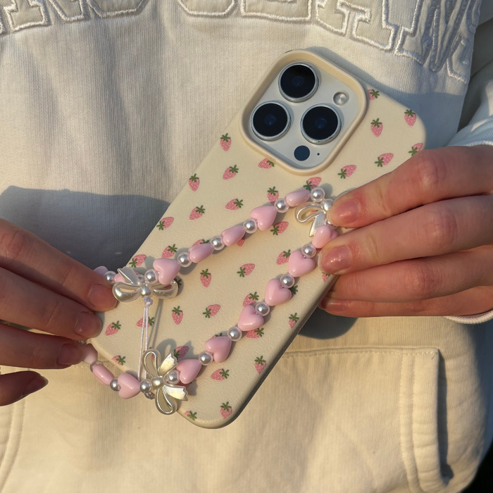 Hand holding a phone with a red strawberry case and Coconut Lane Hearts & Bows beaded strap in sunlight.