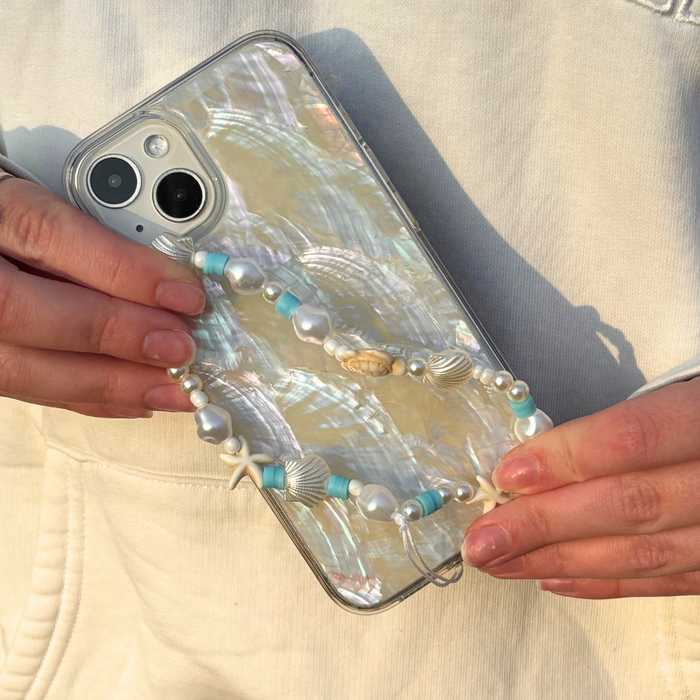 Hands holding a phone in a pearlescent case with a beaded strap decorated with blue and silver ocean-themed charms.