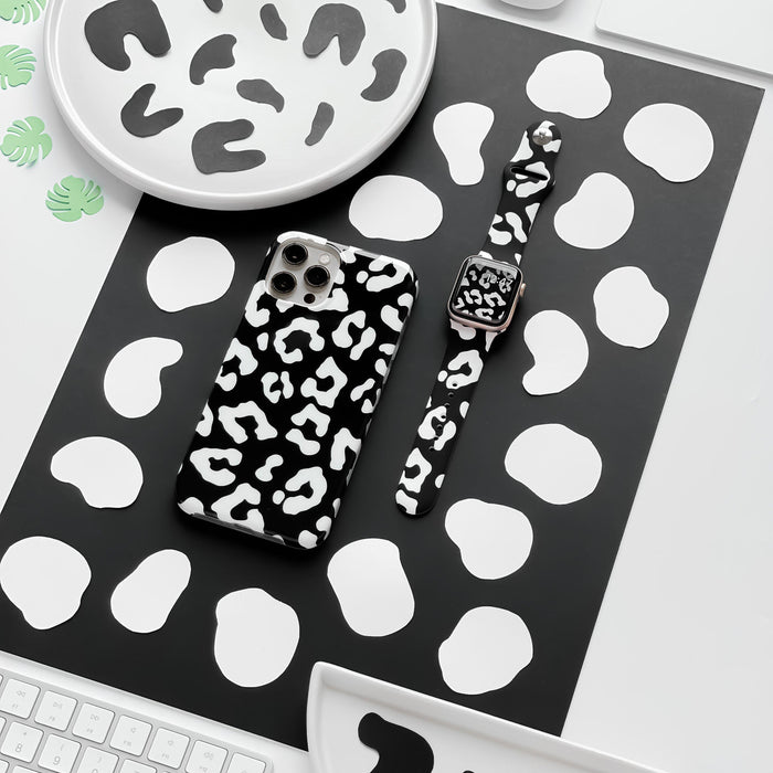 Flat lay of a desk featuring a smartphone with a black and white cow print case and the Black Leopard Apple Watch Strap by Coconut Lane. The desk items rest on a black and white spotted mat, surrounded by plates and coasters with the same black and white pattern.