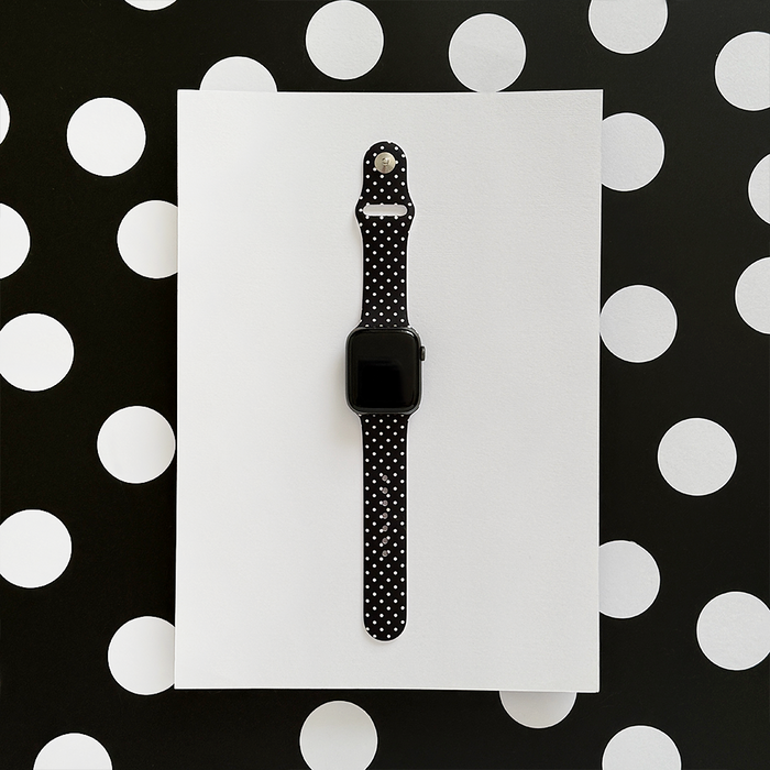 Black polka dot Apple Watch strap by Coconut Lane on a smartwatch, displayed on a white sheet with a polka dot background.