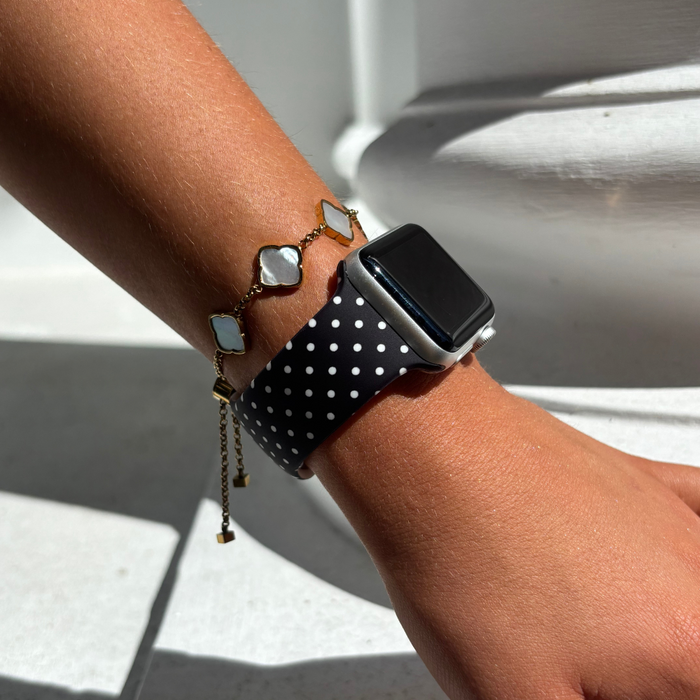 Black polka dot Apple Watch strap and gold bracelet with heart charms on a wrist.