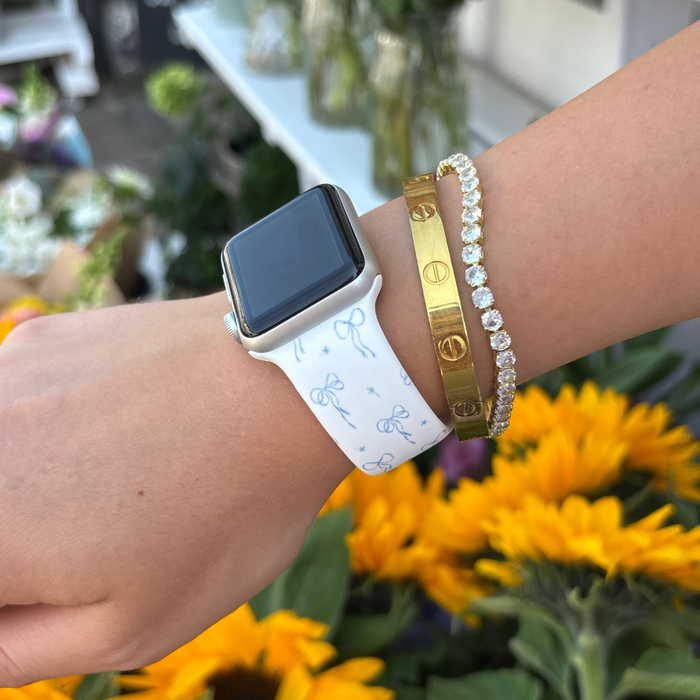 Wrist with a blue bow Apple Watch strap and gold bracelet; sunflowers blurred in the background.