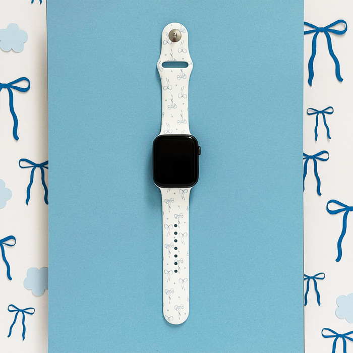 Bluebell Bows Apple Watch strap in pastel blue with bow accents, displayed on a blue background with clouds.