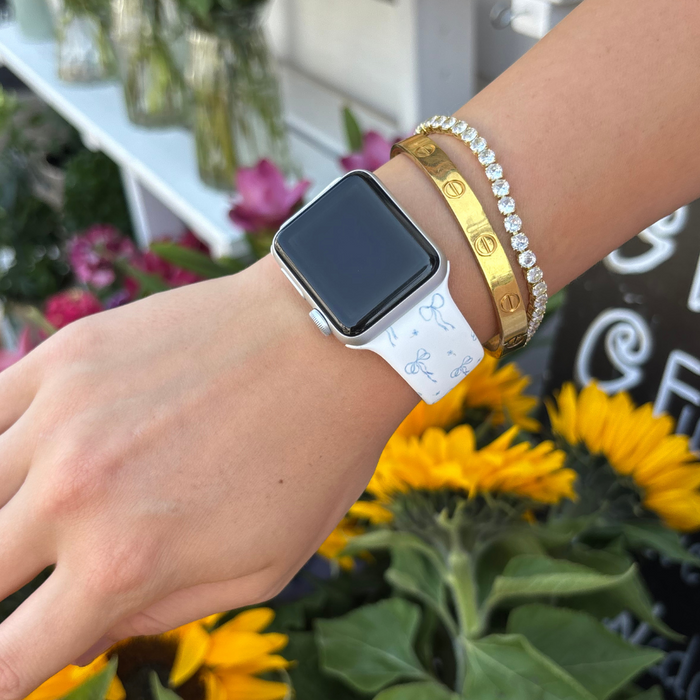Hand wearing blue bow-patterned Apple Watch strap with gold bracelets, flowers blurred in the background.
