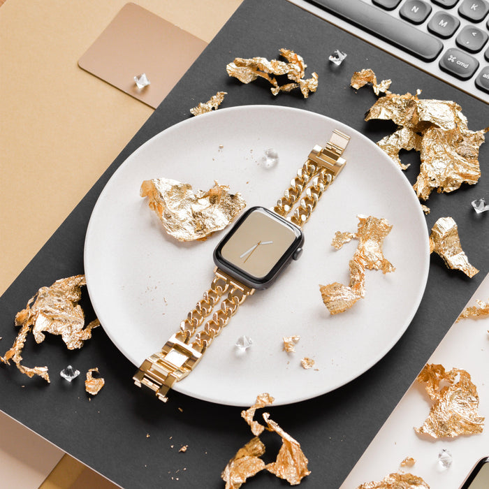 Gold dual-chain Apple Watch strap on a white plate with gold accents, next to a laptop keyboard.