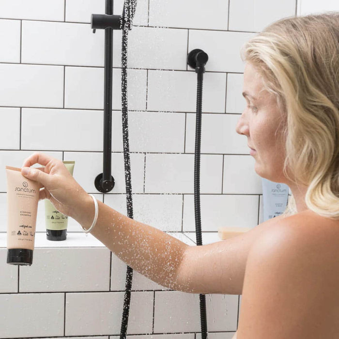 Woman in a shower holding a tube of skincare product with a tiled bathroom background.