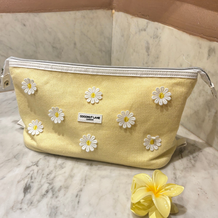 Buttercup yellow expandable makeup bag with embroidered daisies on marble next to a yellow plumeria flower.