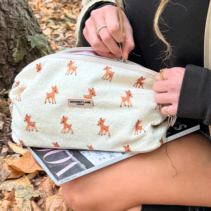 A hand unzips a beige fleecy Coconut Lane expandable makeup bag over a fashion magazine on autumn leaves.
