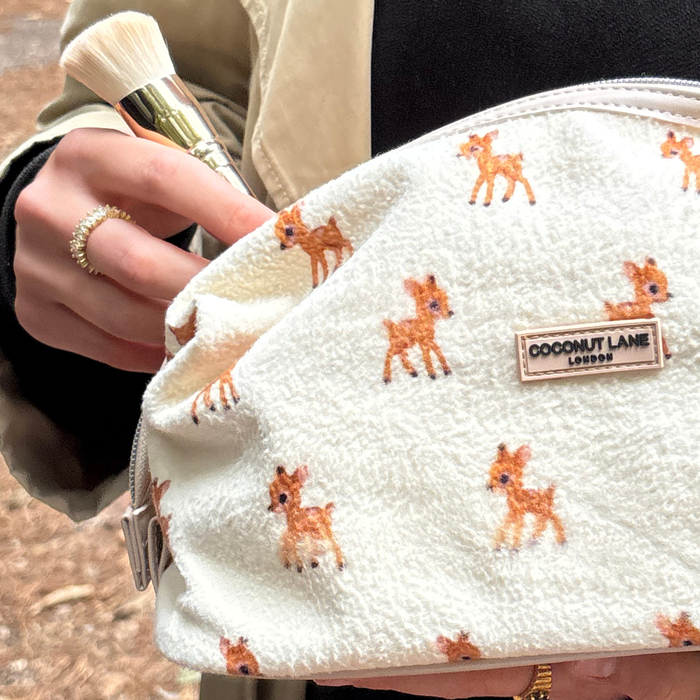 A hand holds a makeup brush and a beige fleecy expandable Coconut Lane makeup bag.