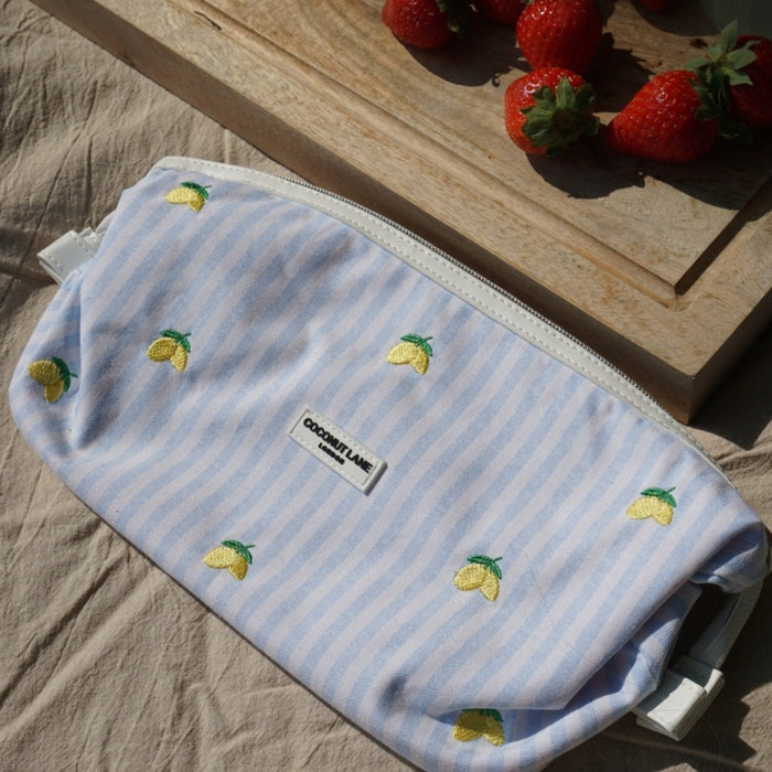 Coconut Lane Living La Vida Lemon yellow expandable makeup bag with lemon print beside strawberries on beige fabric.