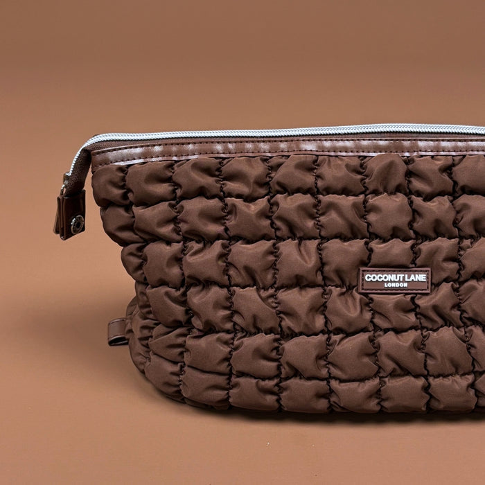 Quilted chocolate brown expandable makeup bag with zipper and label, shown on a brown background.