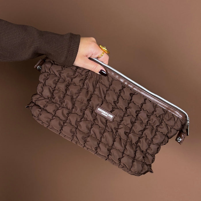Quilted chocolate brown Coconut Lane expandable makeup bag held by a hand in front of a matching brown background.