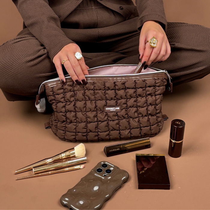 Person cross-legged with a brown quilted makeup bag, beauty products, and phone on a beige surface.
