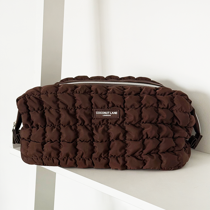 Quilted chocolate brown expandable makeup bag with zipper detail, displayed on a white shelf.