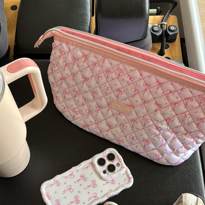 Quilted makeup bag with dainty bow pattern, matching phone case and travel mug, all on a black surface.