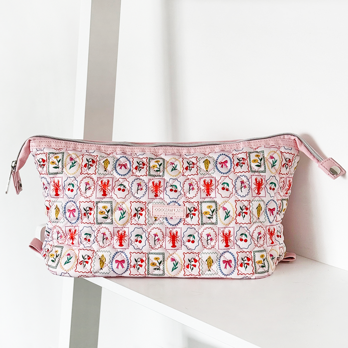 Pink quilted makeup bag with embroidered motifs by Coconut Lane, displayed on a white shelf.