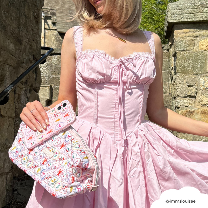 Woman in a light pink dress holding a quilted, patterned Coconut Lane expandable makeup bag and a phone by stone walls.