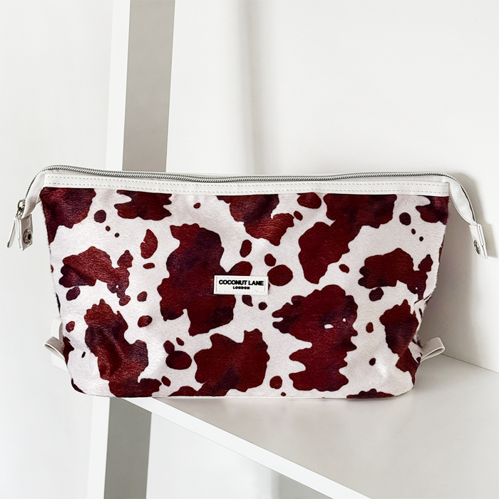 Expandable makeup bag with red and white cow print on a white shelf.