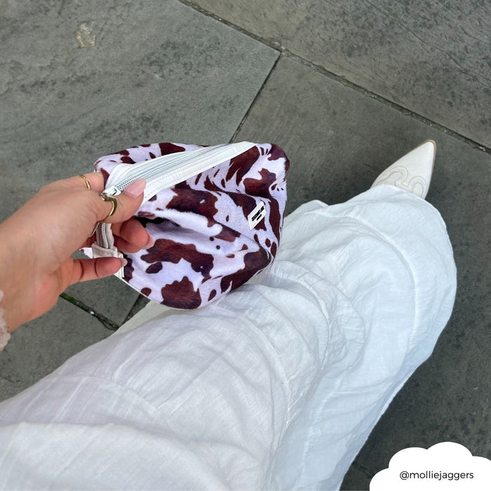 Hand holds a Coconut Lane expandable makeup bag with a Rodeo Cowgirl pattern; person wears white pants on stone pavement.