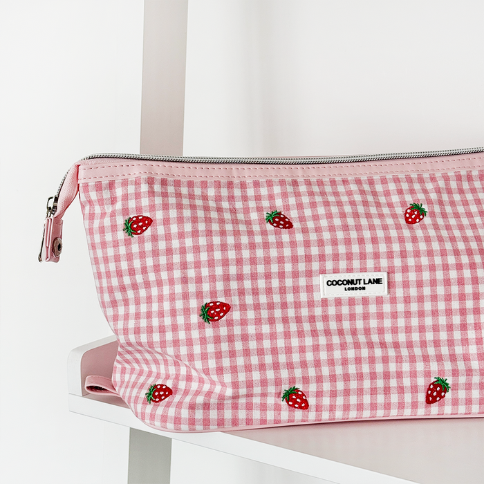 Expandable pink gingham makeup bag with strawberry print on a white shelf.