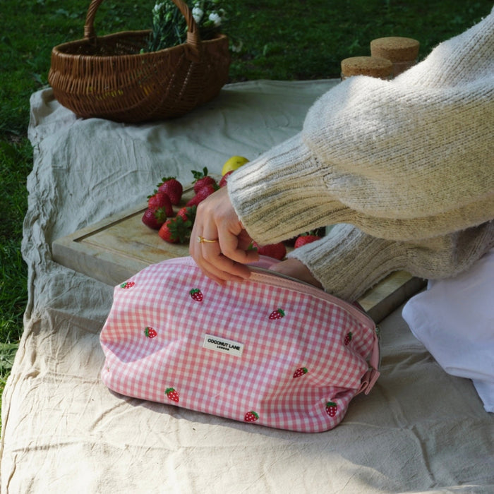 Person opening a pink, strawberry-patterned Coconut Lane expandable makeup bag on a picnic blanket with fruit.