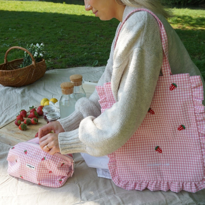 Expandable pink makeup bag with a strawberry pattern being opened on a picnic blanket outdoors.