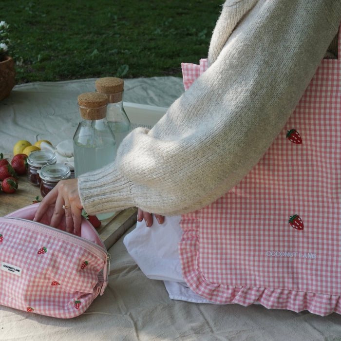 Strawberry Picnic makeup bag by Coconut Lane with pink strawberry print, shown on a picnic blanket with drinks and strawberries.