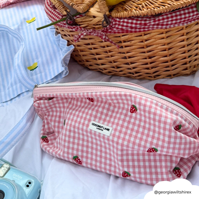 Coconut Lane expandable makeup bag in pink strawberry print on a picnic blanket, surrounded by small accessories.