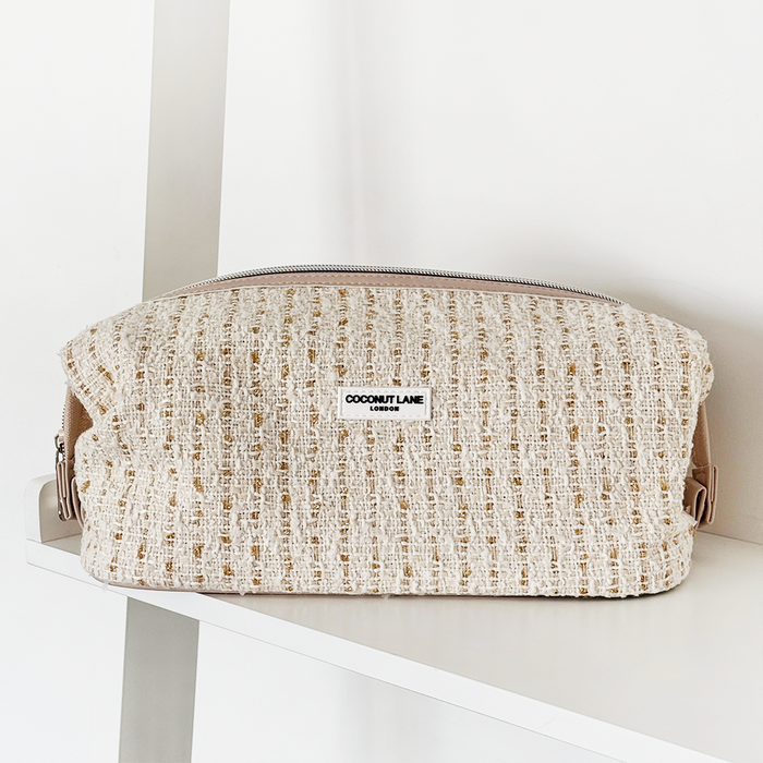 Vanilla textured tweed expandable makeup bag with a silver zip, placed on a white shelf.
