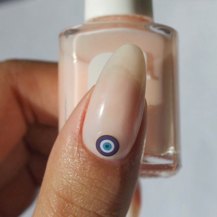 Eye Spy an Evil Eye blue evil eye decal on a sheer nude nail, holding the bottle