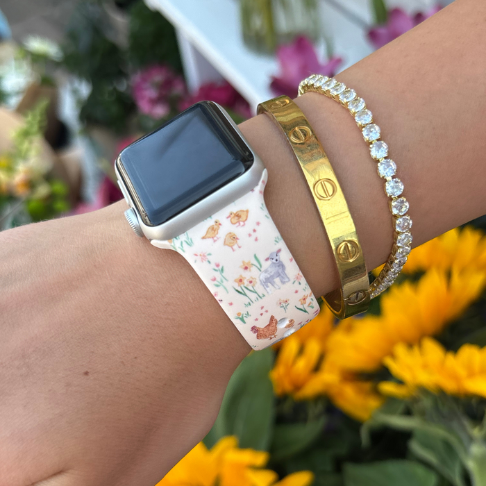 Coconut Lane Farmyard Friends Apple Watch strap on wrist with gold and diamond bracelets, near yellow flowers.