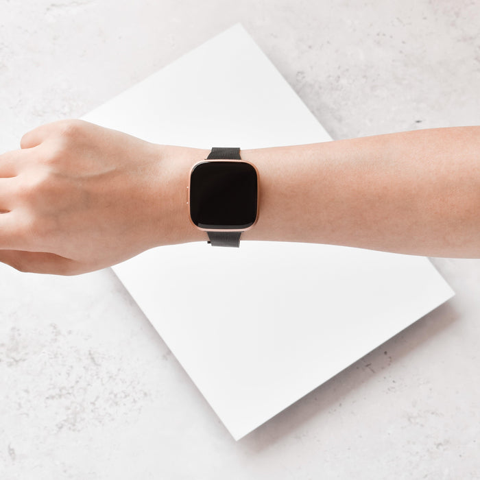Hand wearing a black Slim Milanese Coconut Lane Fitbit strap smartwatch on a white surface beside a blank sheet.