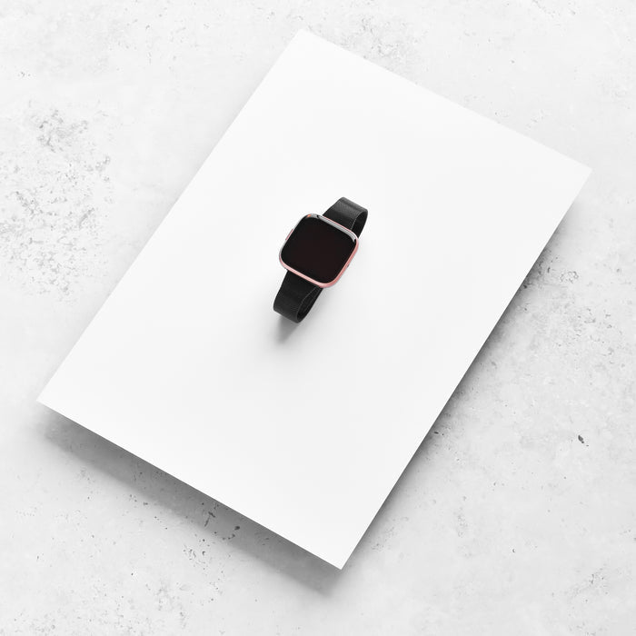 Black smartwatch with a slim black Milanese mesh strap on a white sheet and light gray surface.